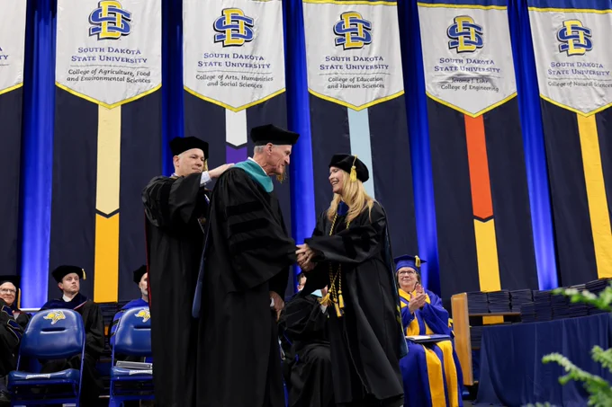 Graduation ceremony at South Dakota State University, featuring professors in academic regalia awarding a hood to Dennis Dougaard on stage. Banners for various colleges, including Agriculture, Food and Environmental Sciences, hang in the background. Audience members and faculty are seated.