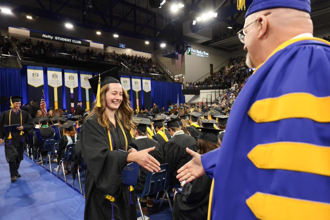 A person in a graduation cap and gown receives a handshake at a commencement ceremony in a large gymnasium. The backdrop features a stage with banners and an audience seated in the bleachers. Visible signage includes "Hefty Student Club."
