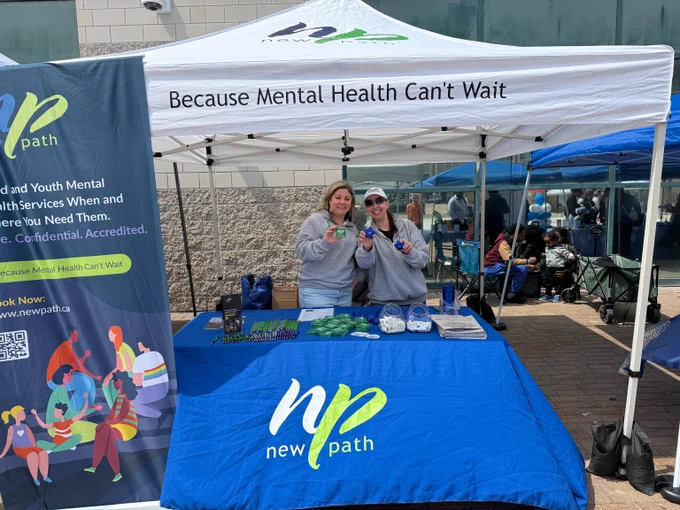 Two women stand behind a New Path mental health awareness booth under a white canopy with the slogan 'Because Mental Health Can't Wait'.