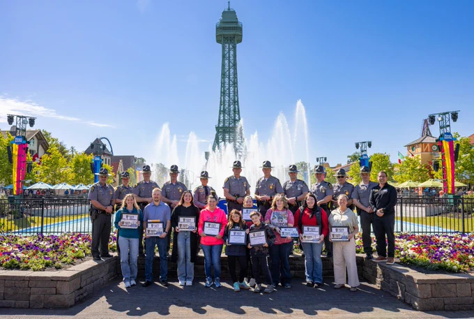 A group of new saved by the belt award winners and Patrol personnel from the Wilmington District, stand in front of a large fountain. In the background is Kings Island amusement park. The winners hold plaques, and colorful flowers adorn the foreground.