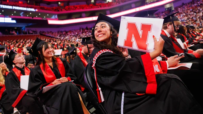A female graduate, seated among other graduates on the floor of Pinnacle Bank Arena, smiles as she holds up her newly earned degree for family in the crowd to see. (Jordan Opp/University Communication and Marketing)