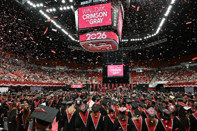 Color indoor photo of crimson and gray confetti falling from the ceiling of Beasley Coliseum while grads look on. Words on a jumbo iron read Win The Day For Crimson and Gray