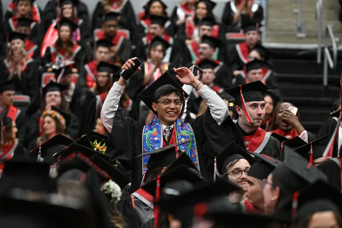 Color indoor photo of a grad raising his arms up above his head in a crowd of other grads. 