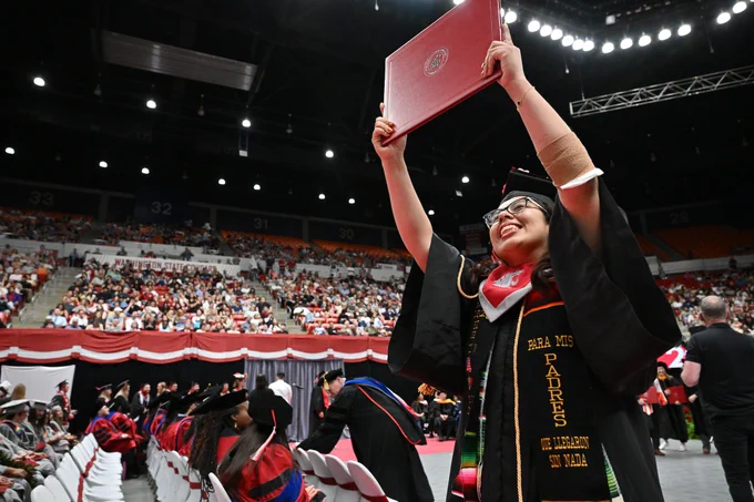 Color indoor photo of a grad walking down an aisle holding a crimson diploma cover above their head. 