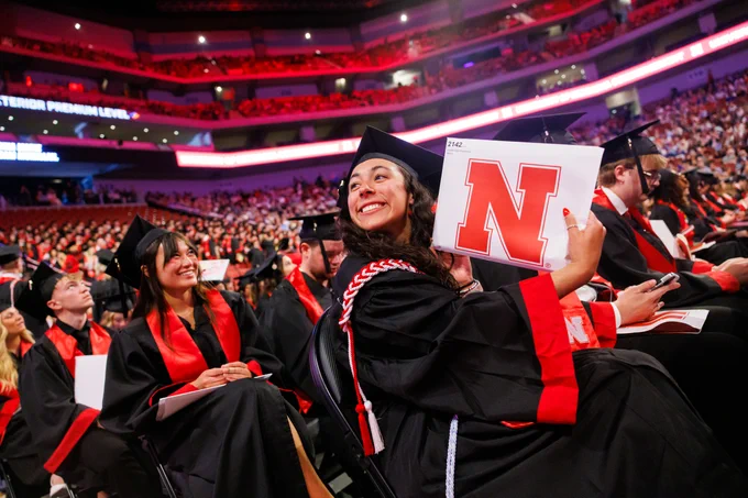 A student holds up her diploma in a white envelope with a red Nebraska N on it as she smiles towards the crowd