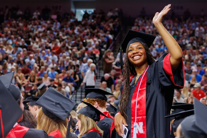 A student smiles and waves to the crowd in graduation regalia 