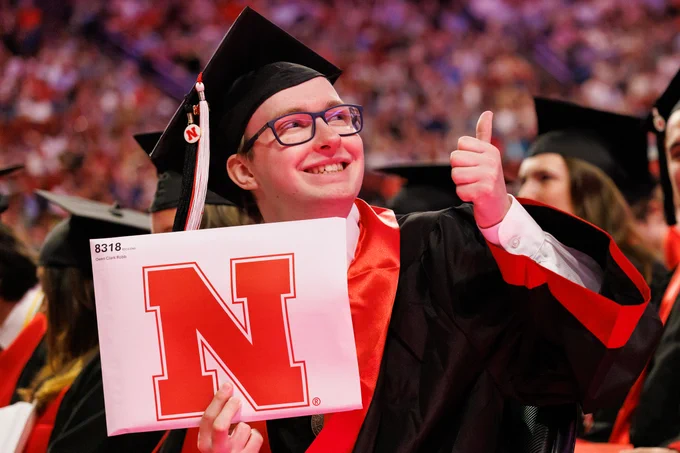 A student holds up his diploma in a white envelope with a red Nebraska N on it as he gives a thumbs up to the crowd