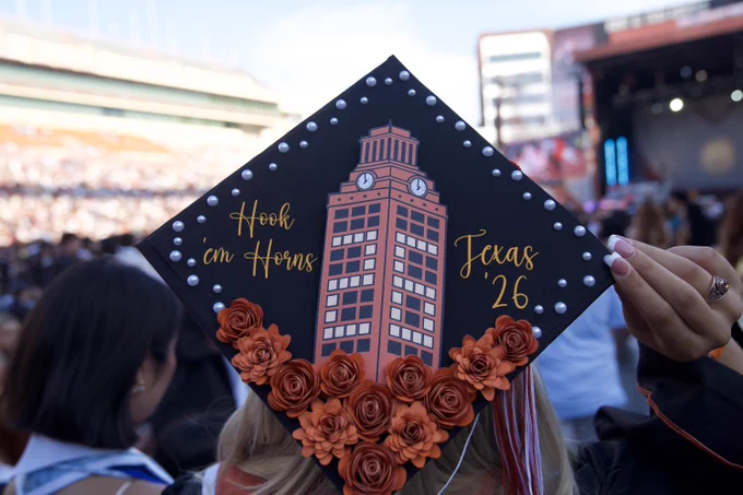 A graduation cap decorated with \"Hook 'em Horns\" text, a building graphic, \"Texas '26\", and orange roses. The cap is black with pearl accents along the edges. A hand holds the cap, with a college ring visible, and a crowd is blurred in the background.