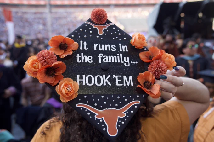A decorated graduation cap reads, \"It runs in the family... HOOK'EM\", adorned with orange flowers and the Texas Longhorns logo. Pearls accentuate the cap's design, held by a hand with teal nail polish.