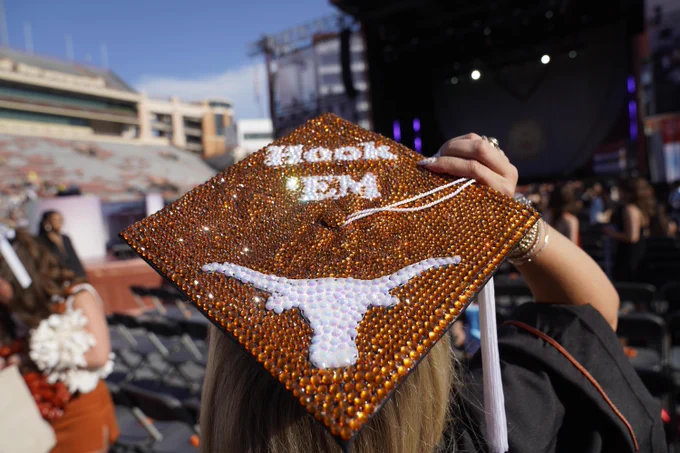 Close-up of a grad cap bedazzled with orange gems and a white longhorn design. The text \"Hook 'Em\" is written above the longhorn. Tassel hangs down, with a stadium in the background.