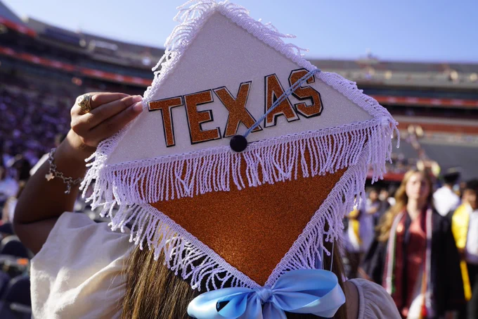 A graduate holds up their decorated mortarboard. The mortarboard is decorated with the word \"TEXAS\" in orange lettering. The graduate is wearing a ring and bracelet with a Texas charm.