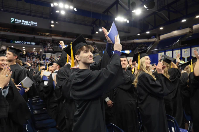 A group of South Dakota State University graduates in black gowns and caps celebrate inside the First Bank & Trust Arena, holding diplomas and clapping. The arena is filled with people and banners.