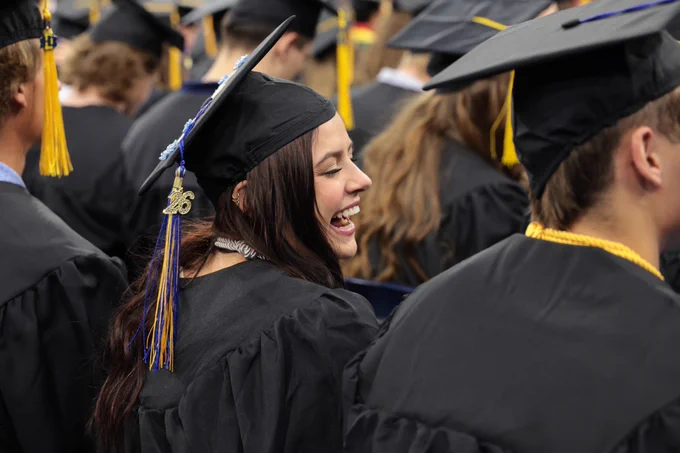 A graduate in a cap and gown smiles joyfully while surrounded by others at a commencement ceremony.