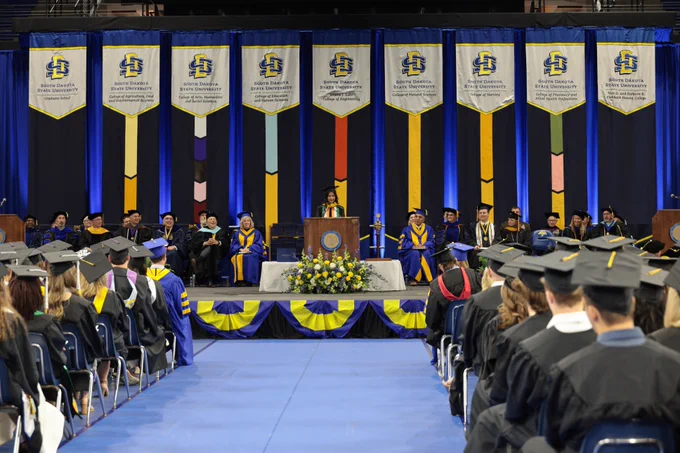 A graduation ceremony is taking place at South Dakota State University. People in academic regalia are seated on a stage adorned with flowers and the university's logo. Attendees in caps and gowns are seated facing the stage. Blue and yellow decorations and banners with the university logo hang in the background.