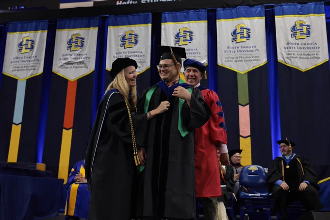 A person in graduation attire is being hooded by two individuals during a ceremony at South Dakota State University. Banners with the university's logo are displayed in the background. Other people in academic regalia are seated on stage.