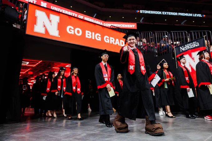A graduate in large Herbie Husker shoes enters the Pinnacle Bank Arena with fellow grads under a display that reads "Go Big Grad"
