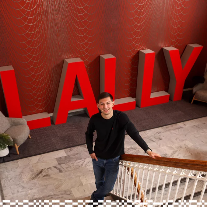 An individual stands on a staircase holding the railing, in front of a large red sign that spells "LALLY." The background features a red wall with geometric patterns.