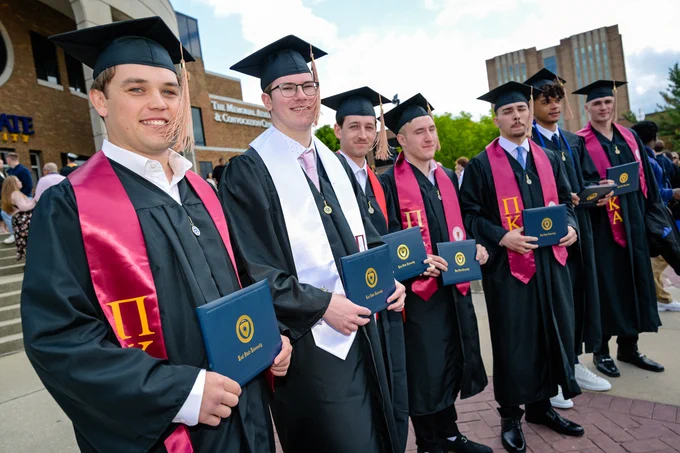 A group of graduates in caps and gowns stand in a row, holding diplomas outside a Kent State's MAC Center. They are wearing red honor cords and smiling towards the camera.