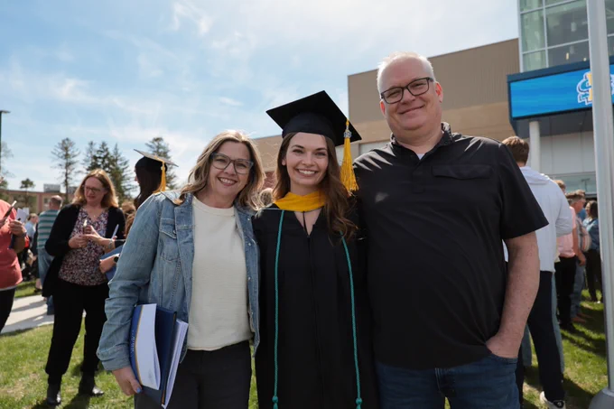 A person in graduation attire stands outdoors between two others who are smiling. There is a grassy area and several people in the background.