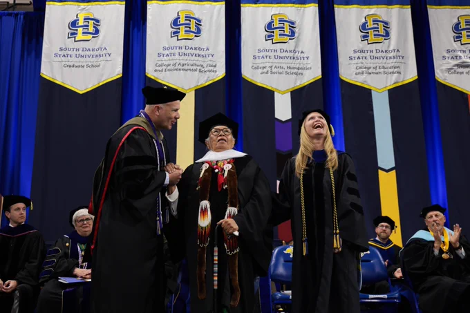 A person in academic regalia is being celebrated at a South Dakota State University event. The backdrop features banners with the university's name and logo. Other individuals in similar attire are seated on stage.