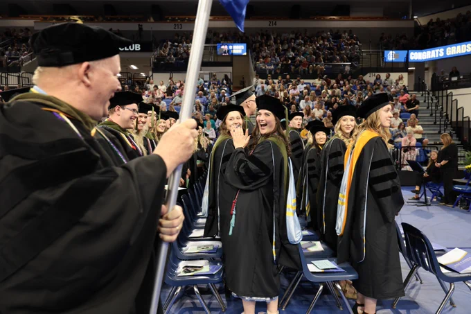 A group of graduates in academic regalia stand and smile at a commencement ceremony in an indoor arena. One person holds a blue flag. The seating area is filled with people, and a banner reads "Congrats Graduates."