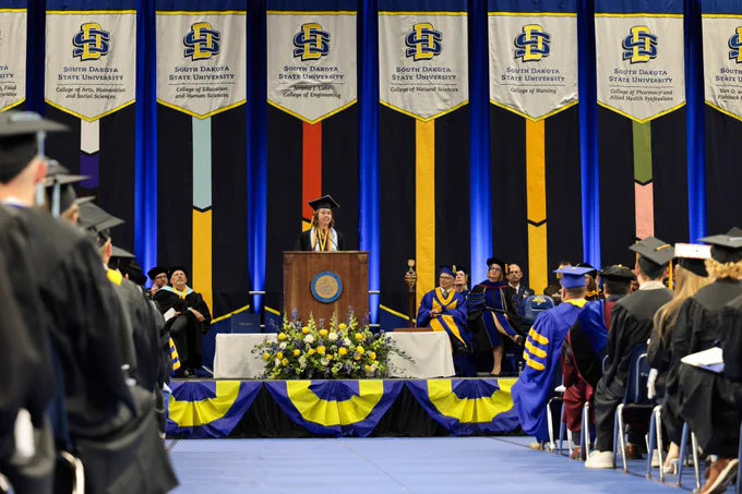 A person stands at a podium on stage during a South Dakota State University graduation ceremony. Academic banners hang above the stage, and the audience consists of graduates in caps and gowns.