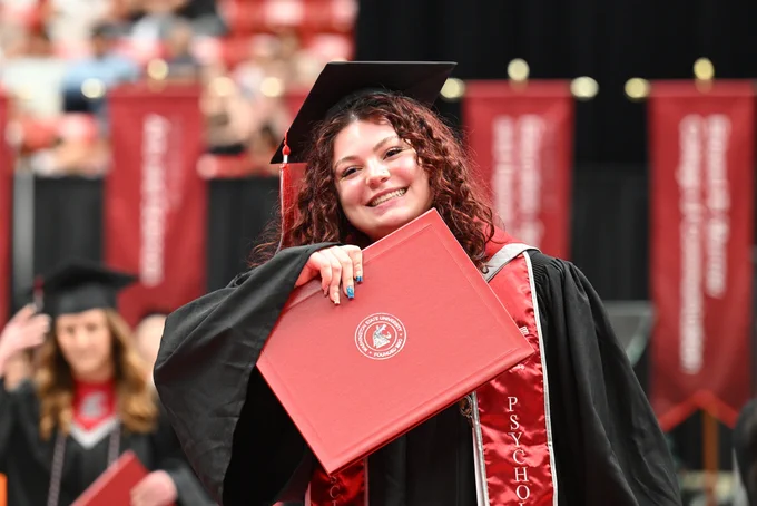 Color indoor photo of a person facing the camera while holding a crimson diploma cover. 