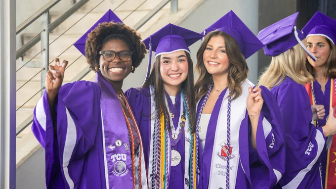 A group of graduates in purple gowns and caps from TCU stand together, smiling and posing. Graduation cords and stoles are visible on their gowns.