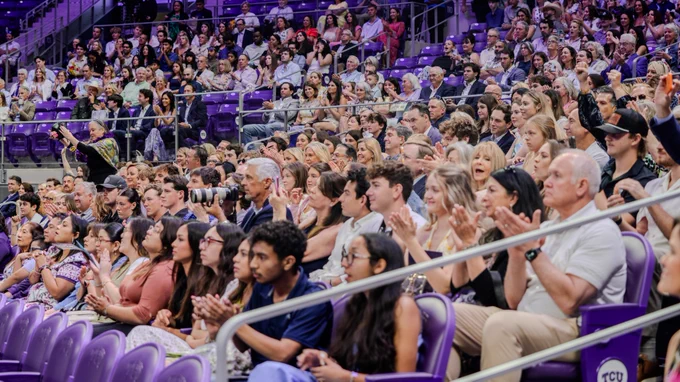 A large audience is seated in a stadium, attentively watching an event. Many individuals are clapping, and the seats are filled with people dressed in various attire.