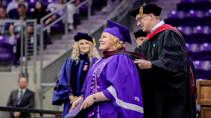 A person in a purple graduation gown and cap smiles as another individual, in academic regalia, places a hood over their shoulders during a ceremony at TCU. The scene takes place in an indoor arena with others in academic attire in the background.