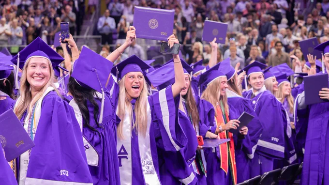 Graduates in purple gowns and caps celebrate at a TCU (Texas Christian University) commencement ceremony, holding diplomas. The scene is lively with many people in the background.