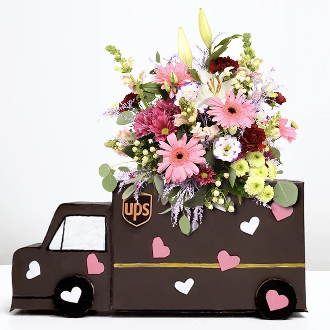 The image shows a UPS delivery truck crafted from cardboard. Atop the cardboard truck is a large, colorful bouquet of pink gerbera daisies, white lilies, yellow blossoms, carnations, and various green foliage.\n\n