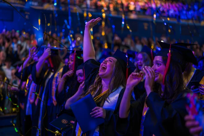 A group of graduates in caps and gowns celebrate with joy and excitement in a crowded auditorium. They are smiling, waving, and holding diplomas as confetti falls around them. The stage and audience are visible in the background, bathed in vibrant blue and purple lighting.