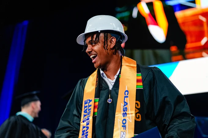 Sure! Here’s an appropriate alt text:

"A graduate wearing a white construction helmet and ceremonial attire smiles at a graduation event. They have a blue gown with a yellow and multicolored stole that reads 'Class of 2026' and 'Kent State University.' Another individual is in the background."