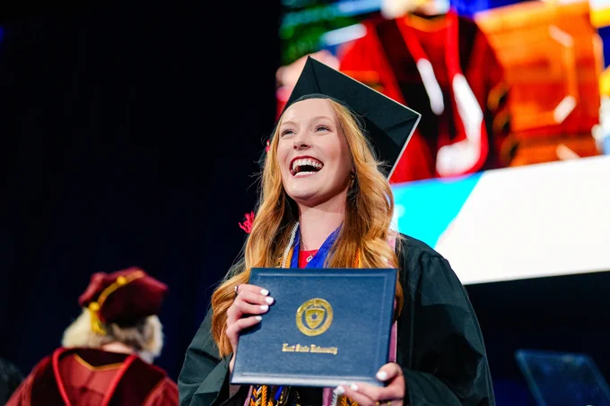 A Kent State graduate smiles broadly while looking upward, holding her diploma on the commencement stage, wearing a red tassel and purple honor cords against a dark background.