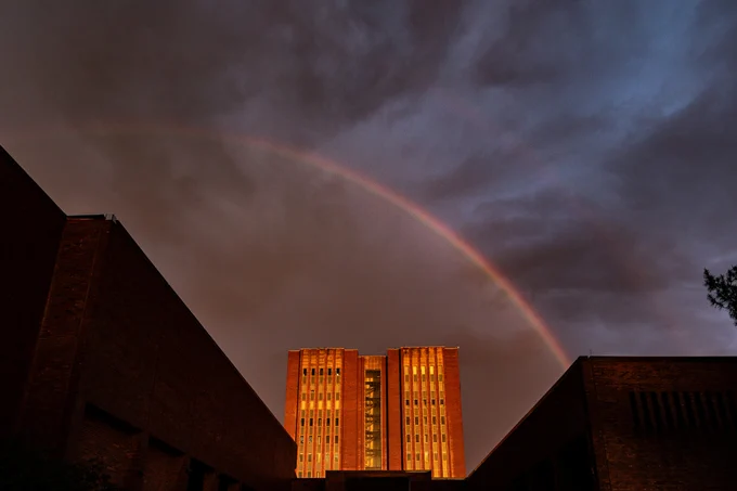 A dramatic sky with a rainbow arches over a tall, illuminated building flanked by two dark structures. The scene is set at dusk with a backdrop of clouds creating a striking contrast.