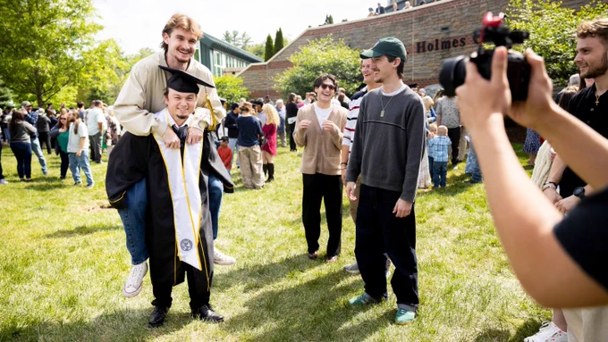 A joyful outdoor graduation scene featuring one person in a cap and gown giving another person a piggyback ride. Friends gather around, smiling and laughing. A "Holmes" building sign is in the background, along with a crowd of people and greenery.