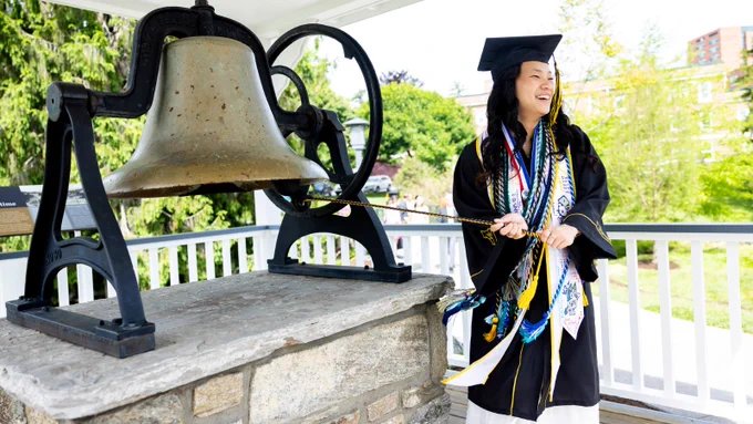 A person in graduation attire stands on a platform, ringing a large bronze bell. The platform is outdoors, surrounded by greenery and a building is visible in the background. The person is wearing multiple colorful tassels and sashes over the gown.