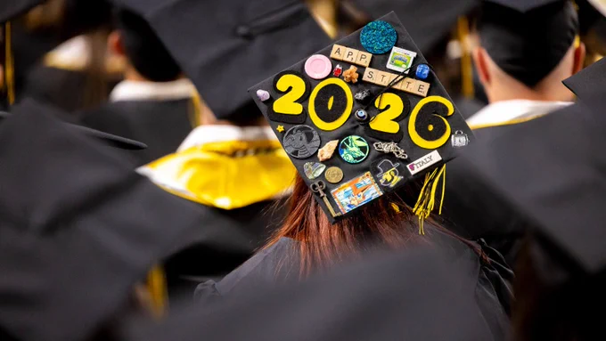 A graduation cap decorated with various items including a space theme and the year "2026" in bold numbers. Students in caps and gowns are seated in rows, a typical graduation setting.