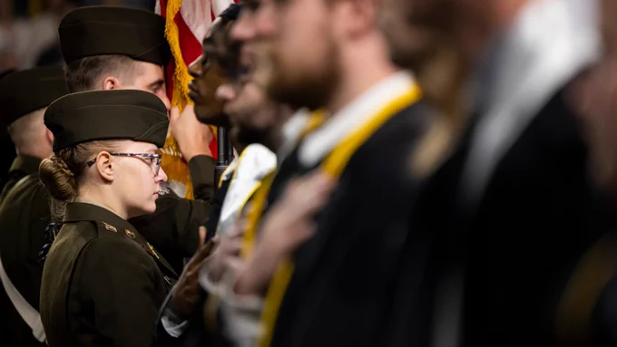 A group of individuals in uniform stand in a row, some with hands over their hearts, facing a flag in the background. The scene appears solemn and formal.