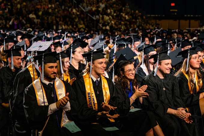 Rows of Kent State University graduates in black caps and gowns applaud during the commencement ceremony, with decorated mortarboards and gold Kent State stoles visible throughout the packed arena.