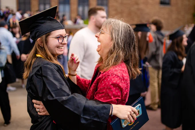 A Kent State graduate in cap and gown laughs joyfully with an older woman in a red floral top, who holds a Kent State diploma folder, as they embrace outside after the commencement ceremony.