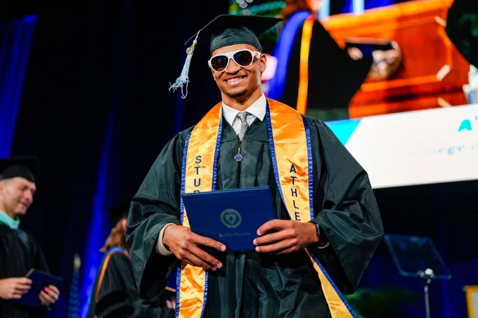 A Kent State student-athlete graduate strides confidently across the commencement stage wearing oversized white sunglasses and a gold student-athlete stole, holding his diploma with a wide smile.