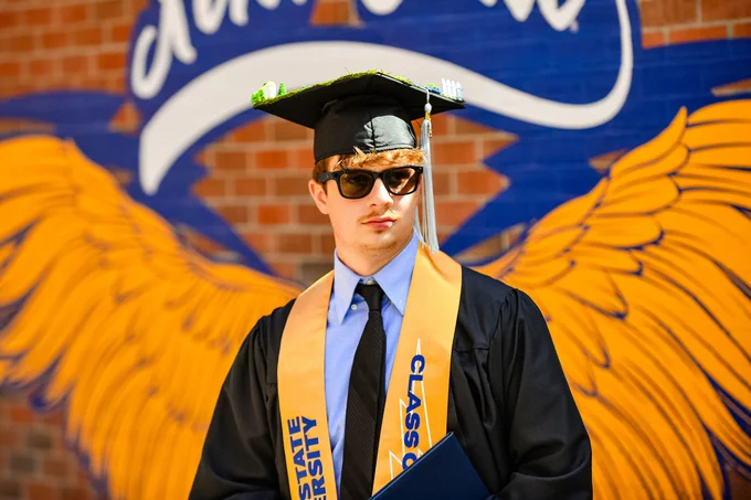 A Kent State graduate poses in sunglasses in front of the golden Flash wings mural on campus, his mortarboard decorated with a miniature landscape scene, diploma in hand.