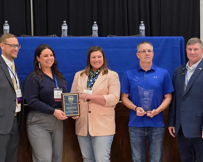 A group of five people stand together for a photo. Two individuals, holding plaques, are in the center. A blue tablecloth with water bottles is in the background.