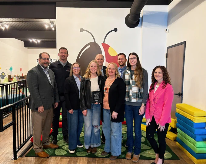 A group of nine people is standing indoors on a colorful rug with a cartoon ladybug mural in the background. They are smiling at the camera, and one person is holding a plaque. The setting appears to be a playful, child-friendly space with bright decor.