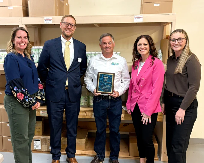 Five people are standing together in front of shelves filled with boxes. One person in the center is holding a plaque. Two individuals are wearing suits and the others are dressed in business casual attire.
