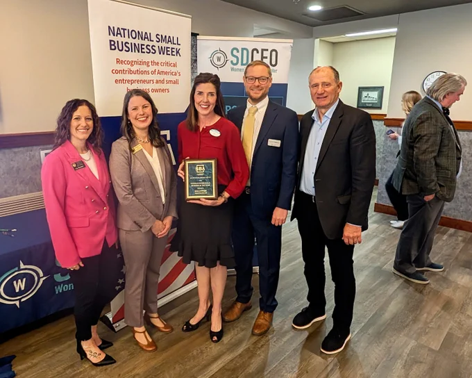 A group of five people are standing indoors, smiling for the camera. The person in the center is holding a plaque. Behind them are banners for "National Small Business Week" and "SDCEO." Other people are in the background.