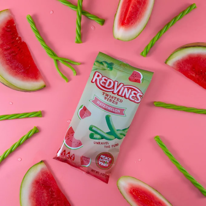 A package of Red Vines Twisted Vines Watermelon candy is surrounded by watermelon slices and loose twisted candies on a pink background.