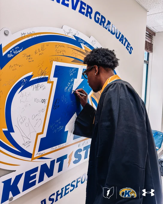Delrecco Gillespie in a graduation gown signs a large Kent State University muralncovered in signatures. The banner includes Kent State's logo and the Under Armour logo.